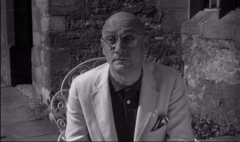 Movie still from “Cul-de-sac” (1966), directed by Roman Polanski – An older man wearing glasses and a jacket; Medium shot, Low angle