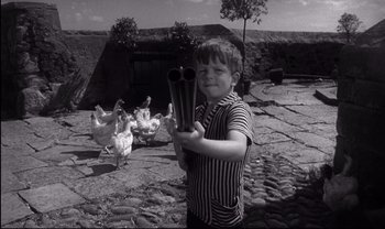 Movie still from “Cul-de-sac” (1966), directed by Roman Polanski – A young boy holding a pair of pipes in front of a group of chickens; Medium shot, High angle