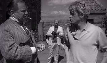 Movie still from “Cul-de-sac” (1966), directed by Roman Polanski – A man smoking a cigarette while two other men watch; Medium shot, Over the shoulder angle