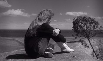Movie still from “Cul-de-sac” (1966), directed by Roman Polanski – A woman sitting on the ground near the ocean; Medium shot, High angle