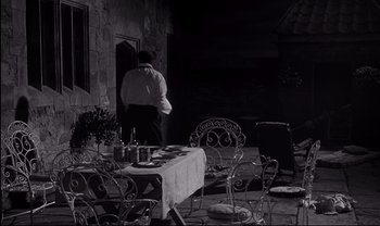 Movie still from “Cul-de-sac” (1966), directed by Roman Polanski – A man standing in front of a table with plates on top of it; Wide shot, High angle