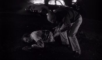 Movie still from “Cul-de-sac” (1966), directed by Roman Polanski – A police officer and a man on the ground in front of a burning car; Medium shot, High angle