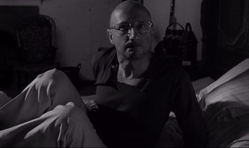 Movie still from “Cul-de-sac” (1966), directed by Roman Polanski – A bald man with glasses sitting on a bed; Medium shot, Low angle