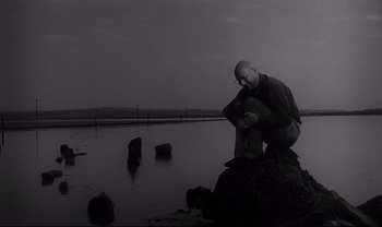Movie still from “Cul-de-sac” (1966), directed by Roman Polanski – A man sitting on top of a rock near a body of water; Wide shot, Low angle