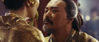 Movie still from “Curse of the Golden Flower” (2006), directed by Yimou Zhang – A man with a beard and a mustache is looking at a doll; Close Up shot, Over the shoulder angle