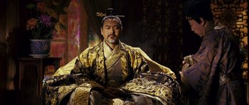 Movie still from “Curse of the Golden Flower” (2006), directed by Yimou Zhang – A man in a gold and black outfit sitting down; Medium shot, Low angle