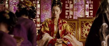 Movie still from “Curse of the Golden Flower” (2006), directed by Yimou Zhang – A woman in a red and gold dress sitting on a chair; Medium shot, Over the shoulder angle