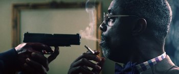 Movie still from “Cut Throat City” (2020), directed by RZA – A man smoking a cigarette while holding a gun pointed at him; Close Up shot, Low angle