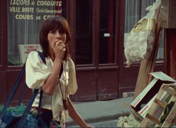 Movie still from “Celine and Julie Go Boating” (1974), directed by Jacques Rivette – A woman standing on the side of the street eating food; Medium shot, Over the shoulder angle