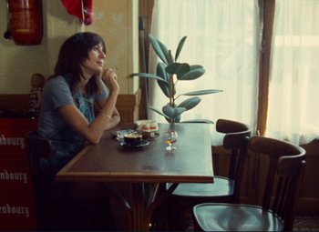 Movie still from “Celine and Julie Go Boating” (1974), directed by Jacques Rivette – A woman sitting at a table with a plate of food; Medium shot, Over the shoulder angle