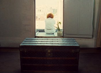 Movie still from “Celine and Julie Go Boating” (1974), directed by Jacques Rivette – A woman sitting on top of a trunk looking out a window; Wide shot, High angle