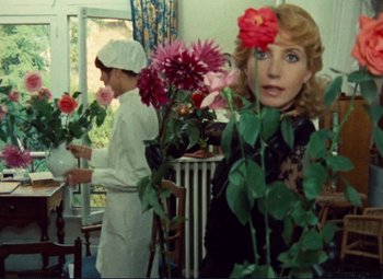 Movie still from “Celine and Julie Go Boating” (1974), directed by Jacques Rivette – Two women are standing in a room with flowers; Medium shot, Over the shoulder angle