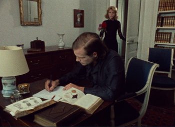 Movie still from “Celine and Julie Go Boating” (1974), directed by Jacques Rivette – A man sitting at a desk writing on a piece of paper next to a woman; Medium shot, Over the shoulder angle