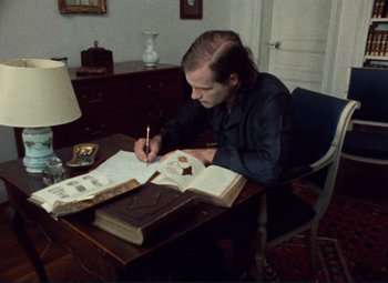 Movie still from “Celine and Julie Go Boating” (1974), directed by Jacques Rivette – A man sitting at a desk writing on a book; Medium shot, High angle