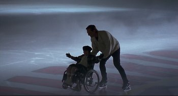 Movie still from “D3: The Mighty Ducks” (1996), directed by Robert Lieberman – A man pushing a boy in a wheel chair on a rink; Wide shot, High angle