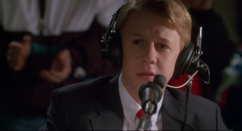 Movie still from “D3: The Mighty Ducks” (1996), directed by Robert Lieberman – A man in a suit and tie wearing a microphone; Close Up shot, Low angle