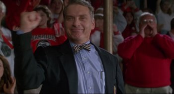 Movie still from “D3: The Mighty Ducks” (1996), directed by Robert Lieberman – A man in a suit and tie smiling for the camera; Medium shot, Low angle