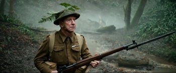 Movie still from “Dad's Army” (2016), directed by Oliver Parker – An old man in uniform holding a rifle in the woods; Medium shot, High angle
