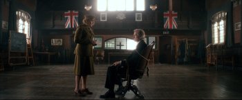 Movie still from “Dad's Army” (2016), directed by Oliver Parker – An old man sitting in a chair next to a woman; Wide shot, Low angle