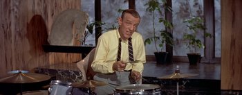 Movie still from “Daddy Long Legs” (1955), directed by Jean Negulesco – A man in a yellow dress shirt and tie playing a drum; Medium shot, High angle