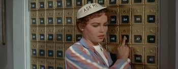Movie still from “Daddy Long Legs” (1955), directed by Jean Negulesco – A woman wearing a hat standing in front of a wall of mailboxes; Close Up shot, High angle