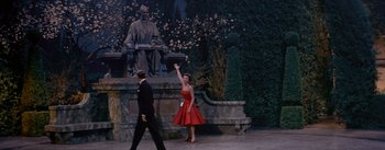 Movie still from “Daddy Long Legs” (1955), directed by Jean Negulesco – A woman in a red dress waves at a man in a black suit; Wide shot, High angle