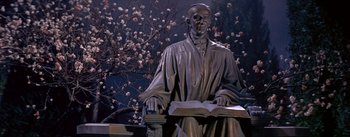 Movie still from “Daddy Long Legs” (1955), directed by Jean Negulesco – A statue of a man sitting on a bench with an open book in his hands; Medium shot, Low angle