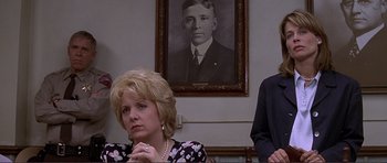 Movie still from “Dante's Peak” (1997), directed by Roger Donaldson – A woman sitting in front of a picture of a man; Medium shot, Over the shoulder angle
