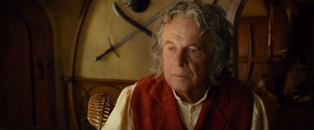 Movie still from “The Hobbit: An Unexpected Journey” (2012), directed by Peter Jackson – An older man in a red vest sitting next to a sword; Close Up shot, Over the shoulder angle