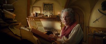 Movie still from “The Hobbit: An Unexpected Journey” (2012), directed by Peter Jackson – An older woman sitting in front of an open book in front of a fireplace; Medium shot, Over the shoulder angle