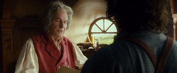 Movie still from “The Hobbit: An Unexpected Journey” (2012), directed by Peter Jackson – An older man wearing a red vest holding papers; Medium shot, Over the shoulder angle