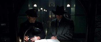 Movie still from “Dark City” (1998), directed by Alex Proyas – Two men in suits and black hats looking at a book; Medium shot, High angle