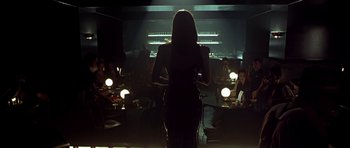 Movie still from “Dark City” (1998), directed by Alex Proyas – A woman standing in front of a group of people in a dark room; Wide shot, Over the shoulder angle