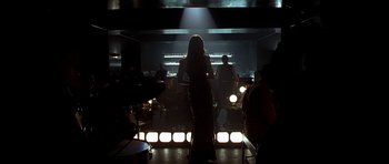 Movie still from “Dark City” (1998), directed by Alex Proyas – A woman standing in front of a group of people in a dark room; Wide shot, Over the shoulder angle