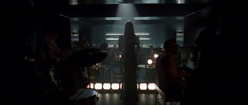 Movie still from “Dark City” (1998), directed by Alex Proyas – A woman standing in front of a group of people in a room; Wide shot, Over the shoulder angle