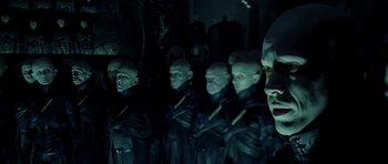 Movie still from “Dark City” (1998), directed by Alex Proyas – A group of men in black suits standing next to each other in a dark room; Medium shot, Over the shoulder angle