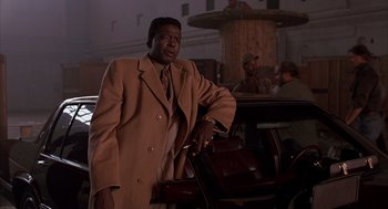 Movie still from “Darkman” (1990), directed by Sam Raimi – A man in a brown coat and a brown tie standing in front of a chair; Medium shot, Low angle