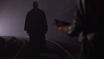 Movie still from “Darkman III: Die Darkman Die” (1996), directed by Bradford May – A man standing in the dark with a gun in the background; Wide shot, Over the shoulder angle