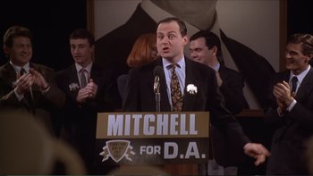 Movie still from “Darkman III: Die Darkman Die” (1996), directed by Bradford May – A man in a suit and tie standing at a podium; Medium shot, High angle