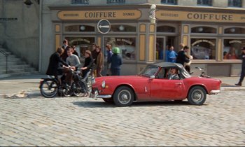 Movie still from “Day for Night” (1973), directed by François Truffaut – A red car driving down a street next to a building; Wide shot, High angle