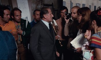 Movie still from “Day for Night” (1973), directed by François Truffaut – A group of people standing around a man holding a flute; Medium shot, Over the shoulder angle