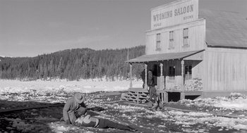 Movie still from “Day of the Outlaw” (1959), directed by André De Toth – Two men are sitting in the snow near a building in the background; Wide shot, High angle
