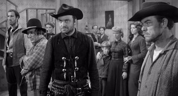 Movie still from “Day of the Outlaw” (1959), directed by André De Toth – A group of people standing in a room with a man in a cowboy hat; Medium shot, Over the shoulder angle