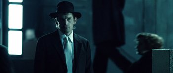 Movie still from “Daybreakers” (2009), directed by Michael Spierig – A man in a suit and hat standing in a dark room; Medium shot, Over the shoulder angle