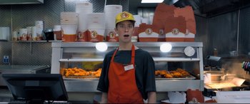 Movie still from “The Kid Who Would Be King” (2019), directed by Joe Cornish – A man wearing an apron and a yellow hat standing in front of a display of hot dogs; Medium shot, Low angle