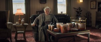 Movie still from “The Kid Who Would Be King” (2019), directed by Joe Cornish – An older man sitting in a chair next to a table with coffee cups on top of it; Wide shot, High angle