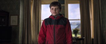 Movie still from “The Kid Who Would Be King” (2019), directed by Joe Cornish – A young boy wearing a red and black jacket in front of a window; Medium shot, Over the shoulder angle