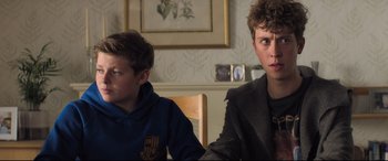 Movie still from “The Kid Who Would Be King” (2019), directed by Joe Cornish – Two young boys sitting at a table in front of a painting; Medium shot, Over the shoulder angle