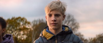 Movie still from “The Kid Who Would Be King” (2019), directed by Joe Cornish – A person wearing a jacket with trees in the background; Close Up shot, Low angle