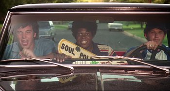 Movie still from “Dazed and Confused” (1993), directed by Richard Linklater – A young man holding a soul pop sign in the back seat of a car; Medium shot, Low angle
